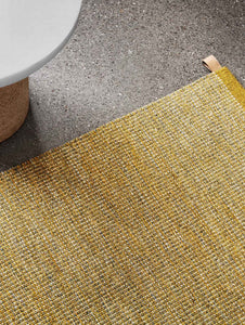 Kasthall’s Golden Ash Harper rug has a gold flecked, tweed-like appearance. This timeless design is flat woven with a thin, low profile, and is made from pure wool.  The rug is shown close up next to a cork side table. 