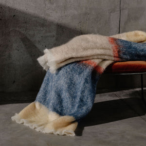 Cushendale's Silare Slate mohair blanket in blue, beige and apricot stripes is folded on a bench and highlighted by sunlight