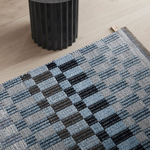 Kasthall chequered Poetry Rug in blues and browns close up in front of wooden stool..