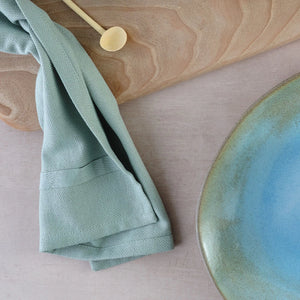 The Organic Company dusty mint cloth napkin on wooden board with artisan ceramic tableware.