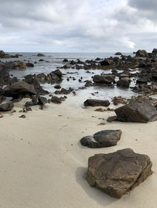 An rocky coastline of Guernsey with a cloudy sky, calm ocean horizon and sandy shore in the foreground. The shoreline is scattered with dark granite rocks across the tideline. 