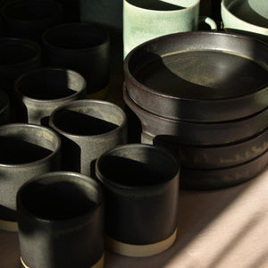 Collection of black tapas dishes and stoneware tumblers on table in sunlight and shadows. Hand-thrown artisan ceramic tableware handmade by Carla Murdoch in the UK.