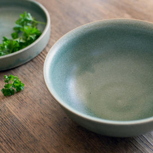 Deep ramen bowl with a softly curved rim shown from above angle, next to a lichen tapas dish with fresh herbs in it. Hand-thrown artisan ceramic tableware handmade by Carla Murdoch in the UK. The bowl has an earthy green lichen colour glaze.