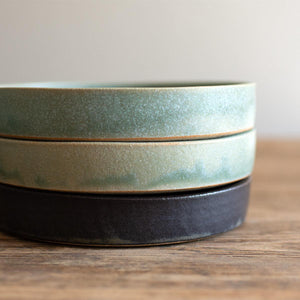Stack of three hand-thrown artisan tapas dishes. Ceramic tableware handmade by Carla Murdoch in the UK. The top two tapas dishes have an earthy green lichen colour glaze. The third tapas dish has a dramatic black glaze with golden and khaki tones.