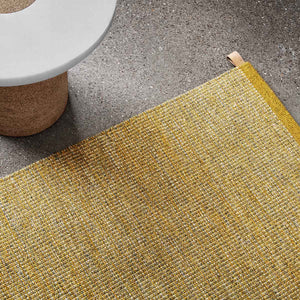 Kasthall’s Golden Ash Harper rug has a gold flecked, tweed-like appearance. This timeless design is flat woven with a thin, low profile, and is made from pure wool.  The rug is shown close up next to a cork side table. 