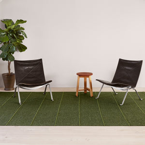 Room set with Kasthall’s Ängsmark Rug, designed by Ilse Crawford. The rug is olive green wool with a lime green stripe. Two black chair and a wooden stool are seated on the rug. A large green houseplant sits behind the rug, again the wall. 