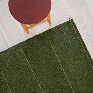 A close up of one end of Kasthall’s Ängsmark Rug, designed by Ilse Crawford. The rug is olive green wool with a lime green stripe, and subtle white flecks running through the weave.  The rug is on a pale wooden floor next to a terracotta wooden stool. 