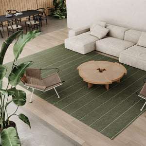 A biophilic designed interior with Kasthall’s Ängsmark Rug, designed by Ilse Crawford, in the centre of the room. The sitting area around the green striped rug has a cream modular sofa, natural woven chairs and a wooden coffee table. Large indoor plants are part of the natural decor. 