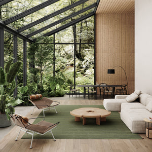 A biophilic (nature-based) designed interior with Kasthall’s Ängsmark Rug, designed by Ilse Crawford, in the centre of the room. This is a large sitting room featuring wooden wall cladding on the far wall and large black framed windows that form the majority of the walls and ceiling, and look out onto lush green woodland. The sitting area around the green rug has a cream modular sofa, natural woven chairs and a wooden coffee table.