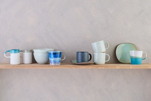 A shelf displaying a selection ceramic mugs, cups and tableware in varying tones of blues and different colour glaze patterns