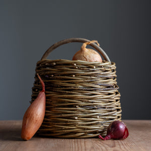 The Mini Forager basket, handwoven by Rachel Bower, has a herringbone weave pattern known as chain wale. The basket is on a wooden table and contains onions. The background is a deep indigo blue colour.