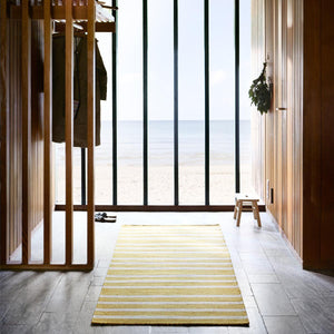 Fablua Living Gold Latte Wool Runner in a wooden clad hallway with a wide window view directly onto a sandy beach and calm blue ocean.
