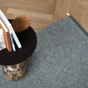 Kasthall’s Amazonite Terrazzo rug is a woven bouclé rug in pure wool and linen. A mix of colours and fibres are woven together to create a wonderfully textured surface. The rug is under a round black and marble side table with books on. The flooring is wood in a herringbone pattern.  