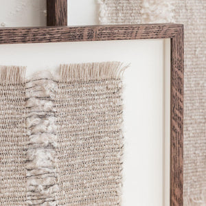 A one-of-a-kind textile artwork woven in mushroom browns, soft white and charcoal tones. Natural fringes add to the delicate and tactile nature of the design. Close up of Oak frame.