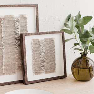 A one-of-a-kind textile artwork woven in mushroom browns, soft white and charcoal tones. Natural fringes add to the delicate and tactile nature of the design. On shelf with brown glass vase containing green foliage.