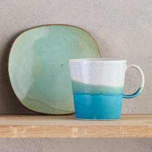The mug features SGW Lab’s signature colour-contrast glaze. Tones transition from speckled grey through to aqua and deep ocean blue., shown on wooden shelf with aqua plate