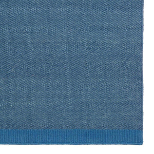 Close up image of textured blue rug with contrasting blue border at end. The weave has a diagonal pattern to it.