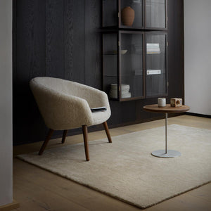 Fabula Living’s Beige Ask Rug is hand-loom pile rug in Gabbeh-dyed New Zealand wool and linen on a cotton warp, shown in a dark room, with a light armchair and side table placed on top of the rug - designed by Jens Landberg Schrøder.