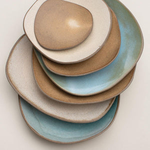 Hana Karim's irregularly-shaped stoneware plates and serving platters stacked together showing a colour palette of blues, beige and honey brown.