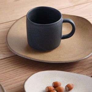 Blue Oda Mug by Julie Damhus Studio is a deep inky blue colour. The mug is shown with cream and brown handmade plates on a wooden table top.