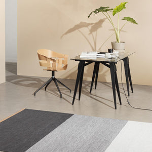 Design House Stockholm Blue/Grey Fields Rug, by Lena Bergström, had a block striped pattern with a light colour stripe one end, graduating to a dark blue grey stripe at the other end. The flatwoven wool rug is positioned in front of a Scandinavian designed desk with plant, and a wooden chair.