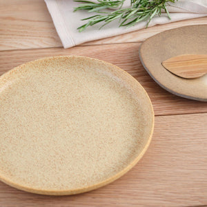 Julie Damhus’ Brown Toto Plate is minimalist in form and decorated with a speckled colour glaze. Shown on a wooden table with another handmade plate, wooden cutlery and rosemary garnish.