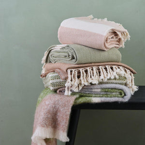 selection of Lüks blankets in pale pinks, moss greens and cream, shown against a green wall. 