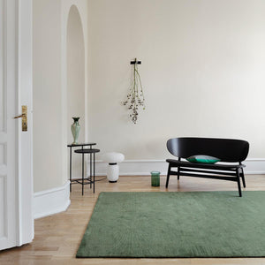 The Fabula Living’s Dusty Green Loke Rug placed on a wooden floor in a light coloured room, with a black bench seat placed on the edge of the rug- designed by Jens Landberg Schrøder