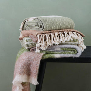 selection of Ferah Blankets, in moss green, pinks and cream. shown against green wall