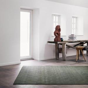 Fabula Living’s Forest Rune Rug, placed on a warm toned dark tiled floor in a light room, behind is a desk with a sculpture & stack of books sat on top and there is a wooden stool beside the desk. - Rug by Jens Landberg Schrøder