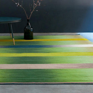 Green Spectrum flatweave wool rug is sat on a white floor with a dark blue wall behind, there is a dark coloured side table and vase sat on the far corner of the rug - by Ptolemy Mann for Gelim