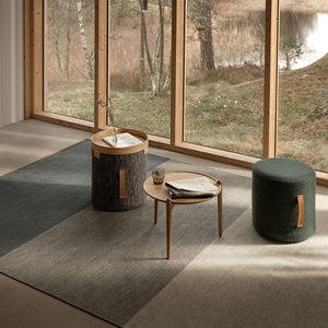 Design House Stockholm Green/Grey Fields Rug, by Lena Bergström, displays block shades of greys and green. Shown in a room with large wooden framed windows over-looking woodland and lake. The rug has side tables placed on top.