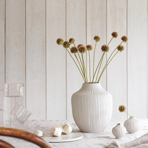 This ceramic vase has a speckled off-white matt finish, with grey lines hand carved into the clay. shown with flowers in vase against wooden white backdrop.