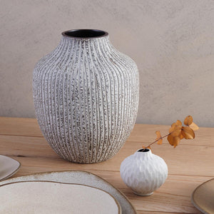 This handmade ceramic vase has a speckled brown texture with a linear grey pattern carved into the clay. Shown on a table with smaller vase and a selection of plates