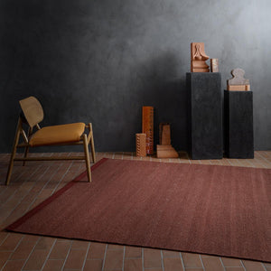 Fabula Livings Lingonberry Una Rug - a deep russet red flatweave rug on red brick floor and under caramel brown leather seated chair. Set against charcoal walls.