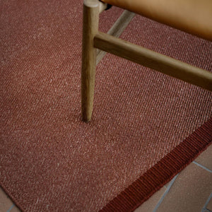 Fabula Livings Lingonberry Una Rug - a deep russet red flatweave rug on red brick floor and under caramel brown leather and wooden chair. 