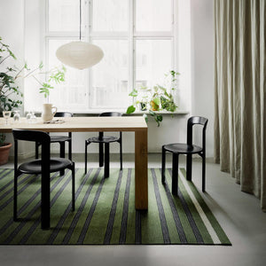 Fabula Living’s Moss/Black Fleur Rug has stripes-within-stripes. The well-balanced pattern is woven in harmonious moss green and black. The rug is placed under a kitchen/dining room wooden table with black chairs. Fresh houseplants add to the biophilic design look of the room. There are large windows of natural light ahead and beige linen curtains hang on the right. 