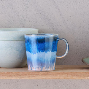 This vibrant blue mug features SGW Lab’s signature colour-contrast glaze. Every handmade mug is unique as the glaze will vary slightly from mug to mug. Shown placed on a wooden shelf sat beside two blue bowls. 