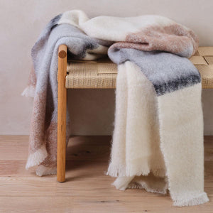 Cushendale's Silare Owl mohair blanket in cream, beige and grey stripes is draped over one end of a wooden bench.