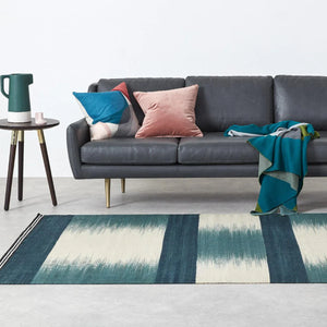 Hand dyed and flatwoven blue rug featuring bold stripes and Ptolemy Mann's signature ikat design. Blue block stripes with ikat stripes. Rug in living room set with grey sofa and side table. 