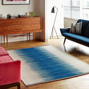 Gelim rug by Ptolemy Mann as a centrepiece in a mid century style living room. The rug has a central ikat stripe in an inky petrol blue which transitions into pale grey in a distinctively ombre style feathered pattern.