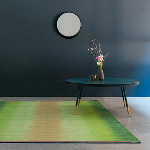 Sage Gelim rug by Ptolemy Mann  in a living room set with dark blue walls and round side table. The ikat stripe rug has bright green tones that graduate gently from vibrant greens to paler tones in the centre of the rug, in a distinctively feathered pattern.