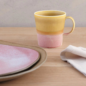 The mug features SGW Lab’s signature colour-contrast glaze. Deep yellow ochre transitions into earthy pink. Every handmade mug is unique as the glaze will vary slightly from mug to mug. Shown placed on a table alongside two stacked plates and cotton napkin.  