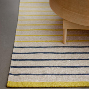 Fabula Living’s Poppy Rug is designed by Lisbet Friis.  A refreshing yellow and beige striped design handwoven in wool and linen. The rug has a fine, tightly woven texture and is also reversible. Close up of the rug under a wooden coffee table. 